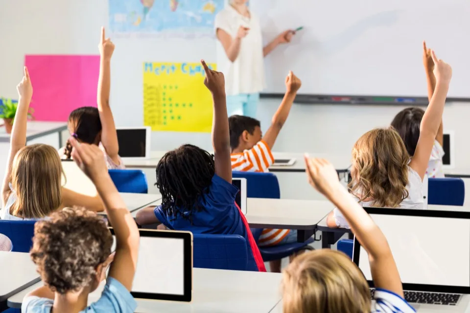Elementary students raising their hands at laptop desks while a teacher leads the lesson from the whiteboard