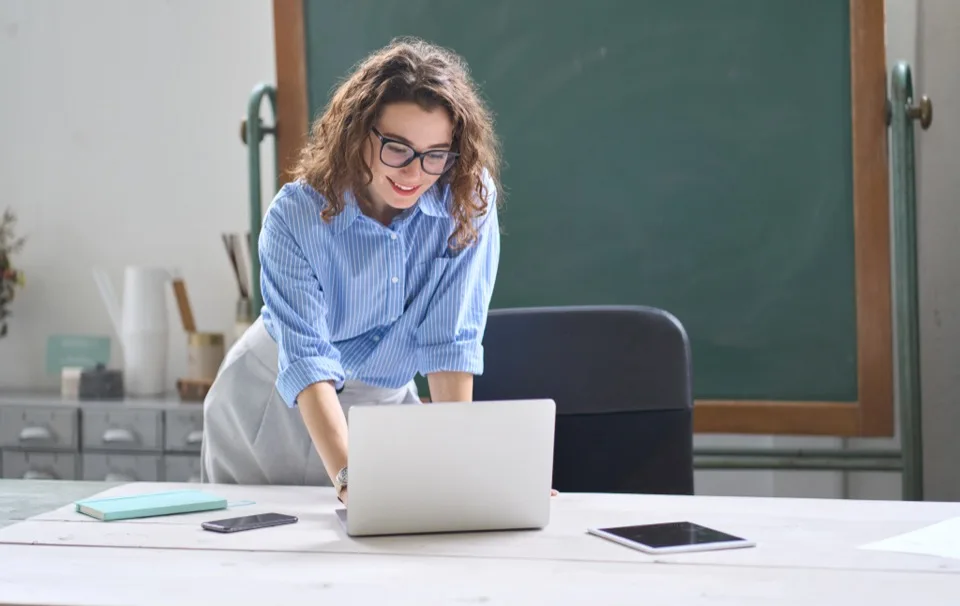 Teacher with curly hair and glasses leaning over a laptop at her desk in front of a green chalkboard