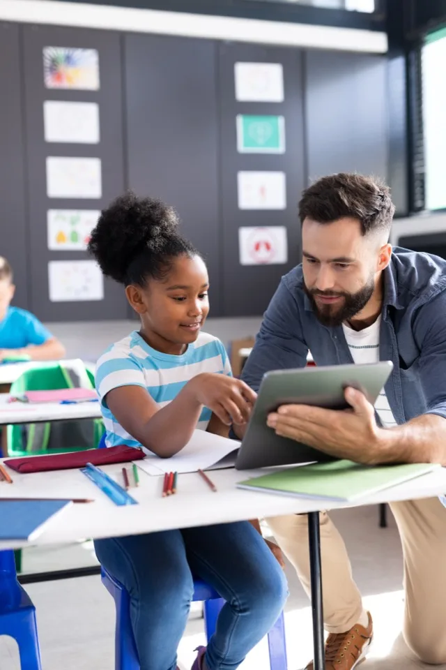 A teacher and a young student looking together at a tablet at a classroom desk